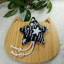 Load image into Gallery viewer, Cat toy made out of black and white star fabric and shaped like a star laying on a wooden board shaped like a cat on a white fuzzy mat with the leaves of a plant in the edge of the photo.
