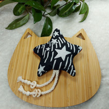 Load image into Gallery viewer, Cat toy made out of black and white star fabric and shaped like a star laying on a wooden board shaped like a cat on a white fuzzy mat with the leaves of a plant in the edge of the photo.