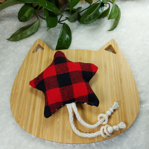 Cat toy made out of red and black buffalo plaid fabric and shaped like a star, laying on a wooden board shaped like a cat. Background is a white fuzzy mat with the leaves of a plant in the edge of the photo.