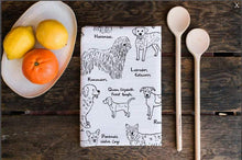 Load image into Gallery viewer, Alphabet Dogs Tea Towel Folded on a table with 2 wooden spoons and a plate of fruit beside it