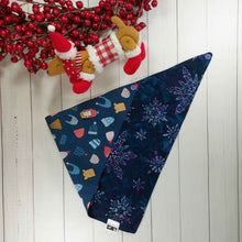 Load image into Gallery viewer,  Dog Bandana made out of blue fabric with snowflakes on one side and winter toques on the other side on it laying flat on a white background with a stuffed toy dog leaning against an artificial red wreath.
