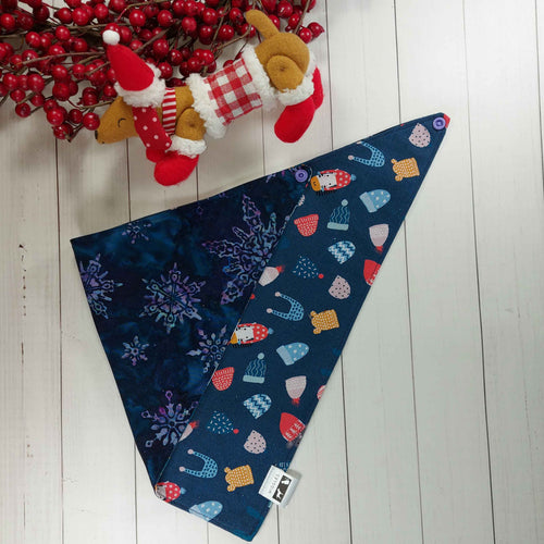  Dog Bandana made out of blue fabric with snowflakes on one side and winter toques on the other side on it laying flat on a white background with a stuffed toy dog leaning against an artificial red wreath.