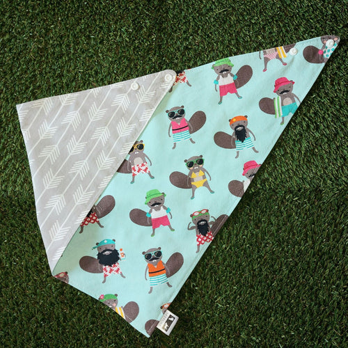 A dog bandana made out of mint green fabric with Beavers dressed in colourful bathing suits all over it laying on artificial turf.