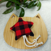 Load image into Gallery viewer, Cat toy made out of red and black buffalo plaid fabric and shaped like a star, laying on a wooden board shaped like a cat. Background is a white fuzzy mat with the leaves of a plant in the edge of the photo.
