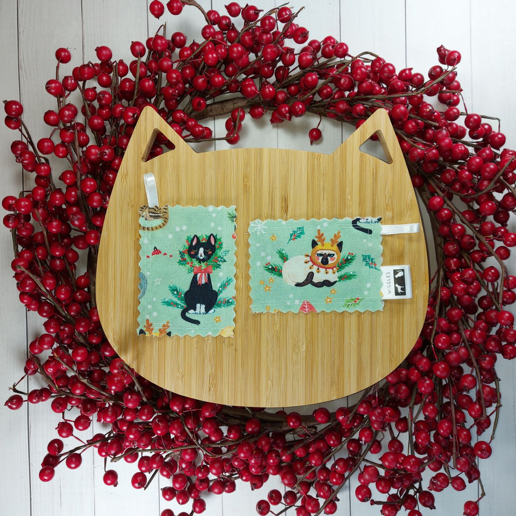 Artificial red berry wreath with cat shaped wooden board in the middle with two rectangular shaped catnip pouches made out of green fabric with cats on them in the middle. 