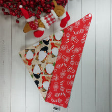 Load image into Gallery viewer, Christmas dog bandana with gnomes on it lying flat next to a small stuffed toy dog leaning against an artificial red Christmas wreath. 
