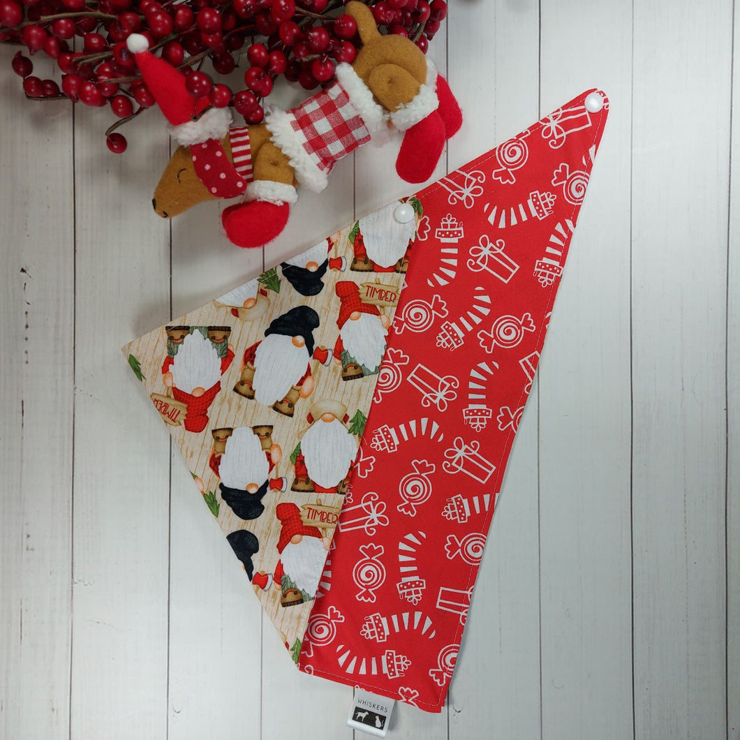 Christmas dog bandana with gnomes on it lying flat next to a small stuffed toy dog leaning against an artificial red Christmas wreath. 