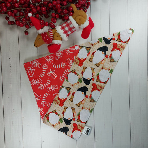 Christmas dog bandana with gnomes on it lying flat next to a small stuffed toy dog leaning against an artificial red Christmas wreath. 