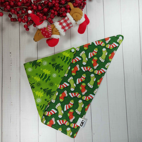 Dog Bandana made out of fabric with green and red Christmas stockings on it laying flat on a white background with a stuffed toy dog leaning against an artificial red wreath.