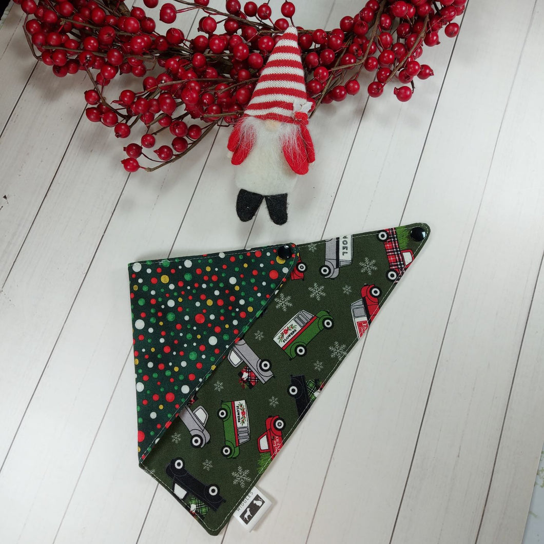 Dog Bandana made out of green fabric with Christmas trucks on one side festive polka dots on the other side on it laying flat on a white background with a stuffed toy dog leaning against an artificial red wreath.