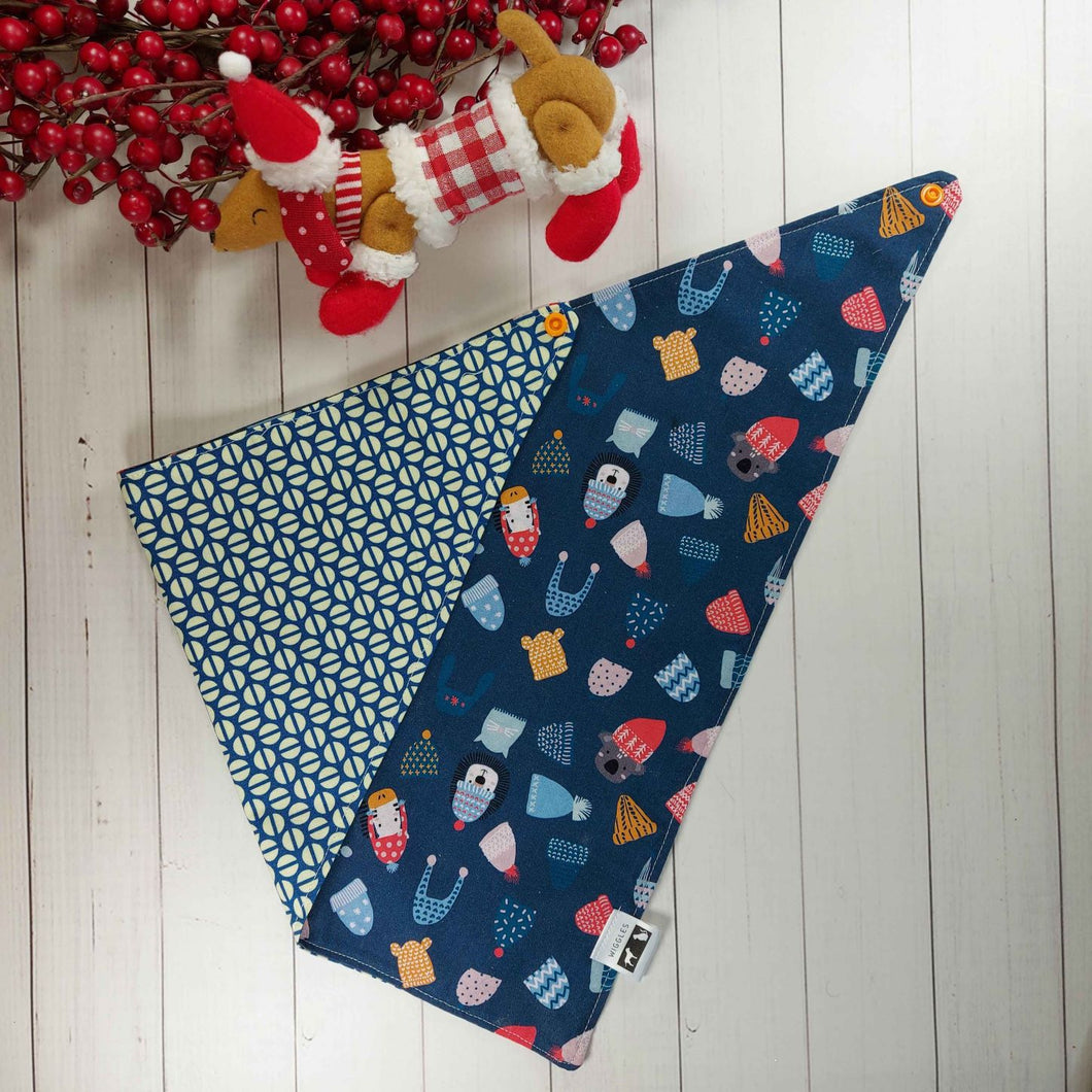 Dog Bandana made out of blue fabric with winter hats on it laying flat on a white background with a stuffed toy dog leaning against an artificial red wreath.