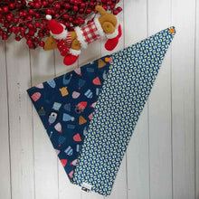Load image into Gallery viewer, Dog Bandana made out of blue fabric with winter hats on it laying flat on a white background with a stuffed toy dog leaning against an artificial red wreath.
